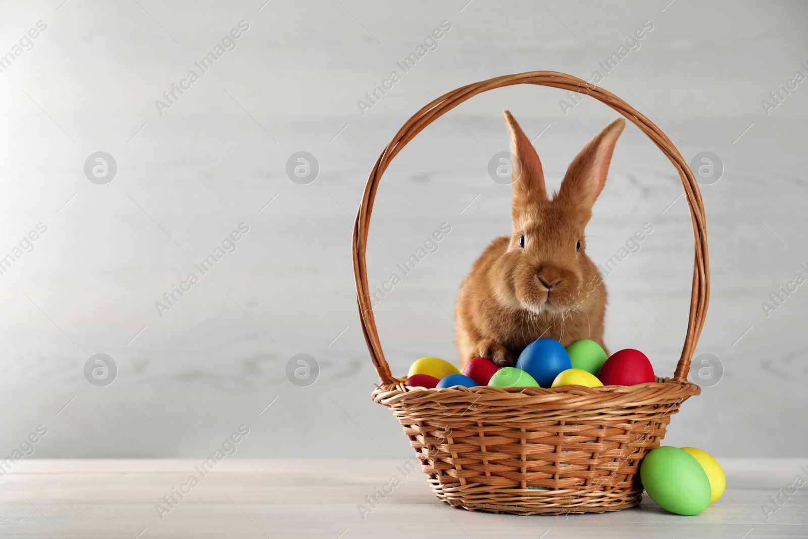 Cute bunny and basket with Easter eggs on white table. Space for text Photo of Cute bunny and basket with Easter eggs on white table. Space for text