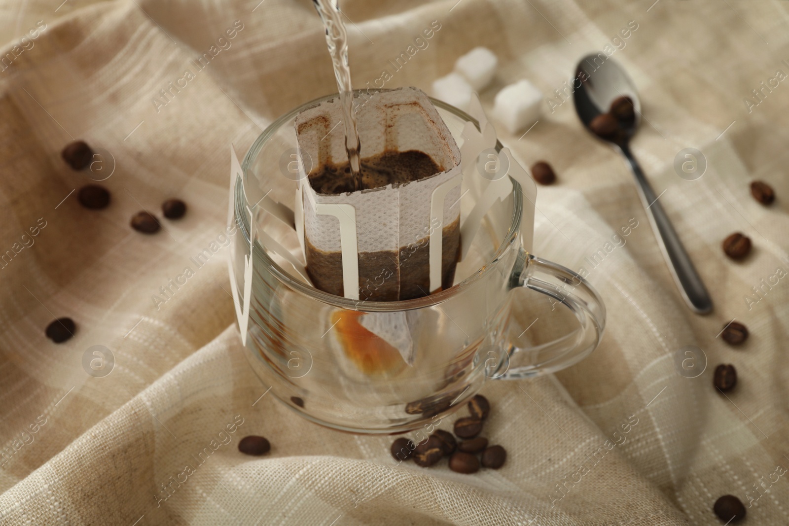Pouring hot water into glass cup with drip coffee bag on beige fabric, closeup Photo of Pouring hot water into glass cup with drip coffee bag on beige fabric, closeup