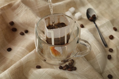 Pouring hot water into glass cup with drip coffee bag on beige fabric, closeup Photo of Pouring hot water into glass cup with drip coffee bag on beige fabric, closeup