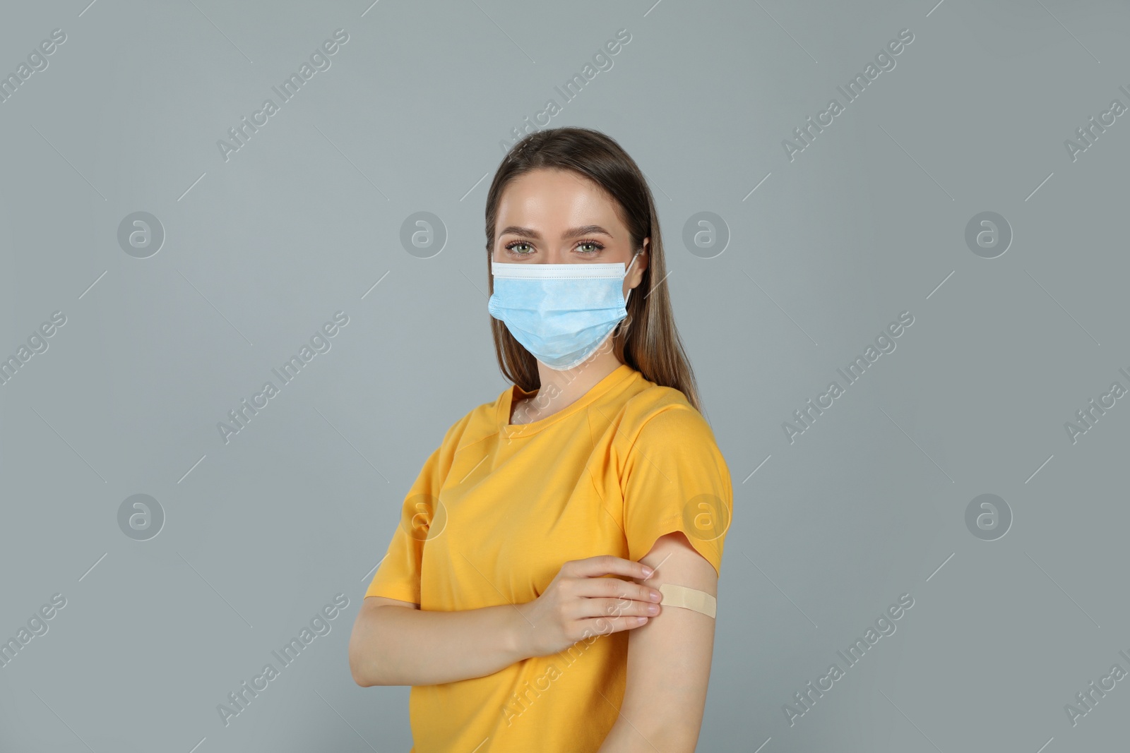 Vaccinated woman with protective mask and medical plaster on her arm against grey background Photo of Vaccinated woman with protective mask and medical plaster on her arm against grey background