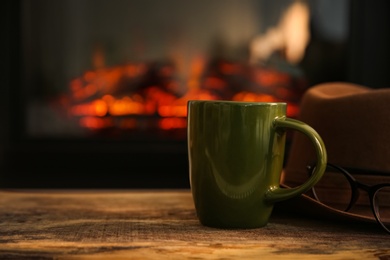 Green cup with hot drink on wooden table against fireplace, space for text Photo of Green cup with hot drink on wooden table against fireplace, space for text