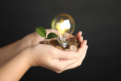 Woman with coins, light bulb and green plant against black background, closeup. Power saving Photo of Woman with coins, light bulb and green plant against black background, closeup. Power saving
