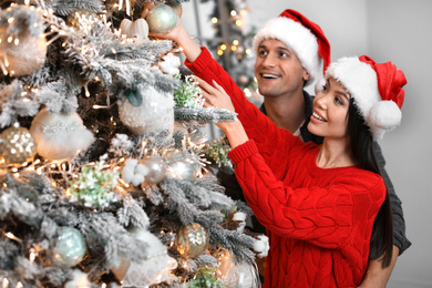 Happy young couple in Santa hats decorating Christmas tree together at home Photo of Happy young couple in Santa hats decorating Christmas tree together at home