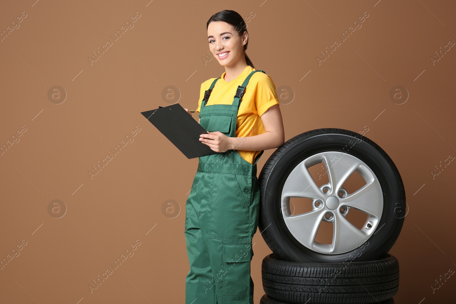 Female mechanic in uniform with car tires and clipboard on color background Photo of Female mechanic in uniform with car tires and clipboard on color background