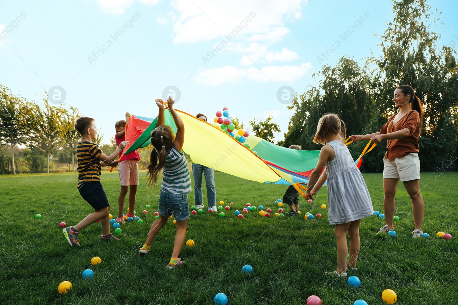 Photo of Group of children and teacher playing with rainbow playground parachute on green grass. Summer camp activity