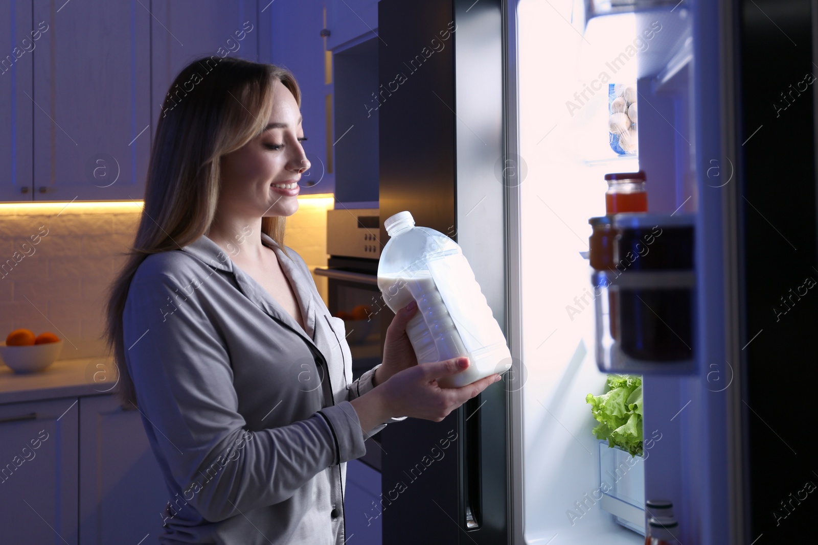 Young woman holding gallon bottle of milk near refrigerator in kitchen at night Photo of Young woman holding gallon bottle of milk near refrigerator in kitchen at night