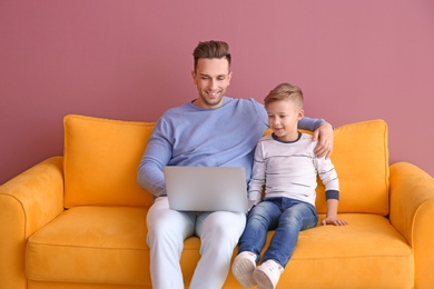 Little boy and his father with laptop sitting on sofa, indoors Photo of Little boy and his father with laptop sitting on sofa, indoors