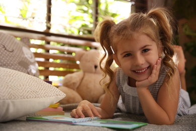 Photo of Cute little girl reading book near window at home