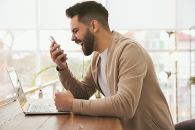 Emotional young man with smartphone at table in office. Online hate concept Photo of Emotional young man with smartphone at table in office. Online hate concept