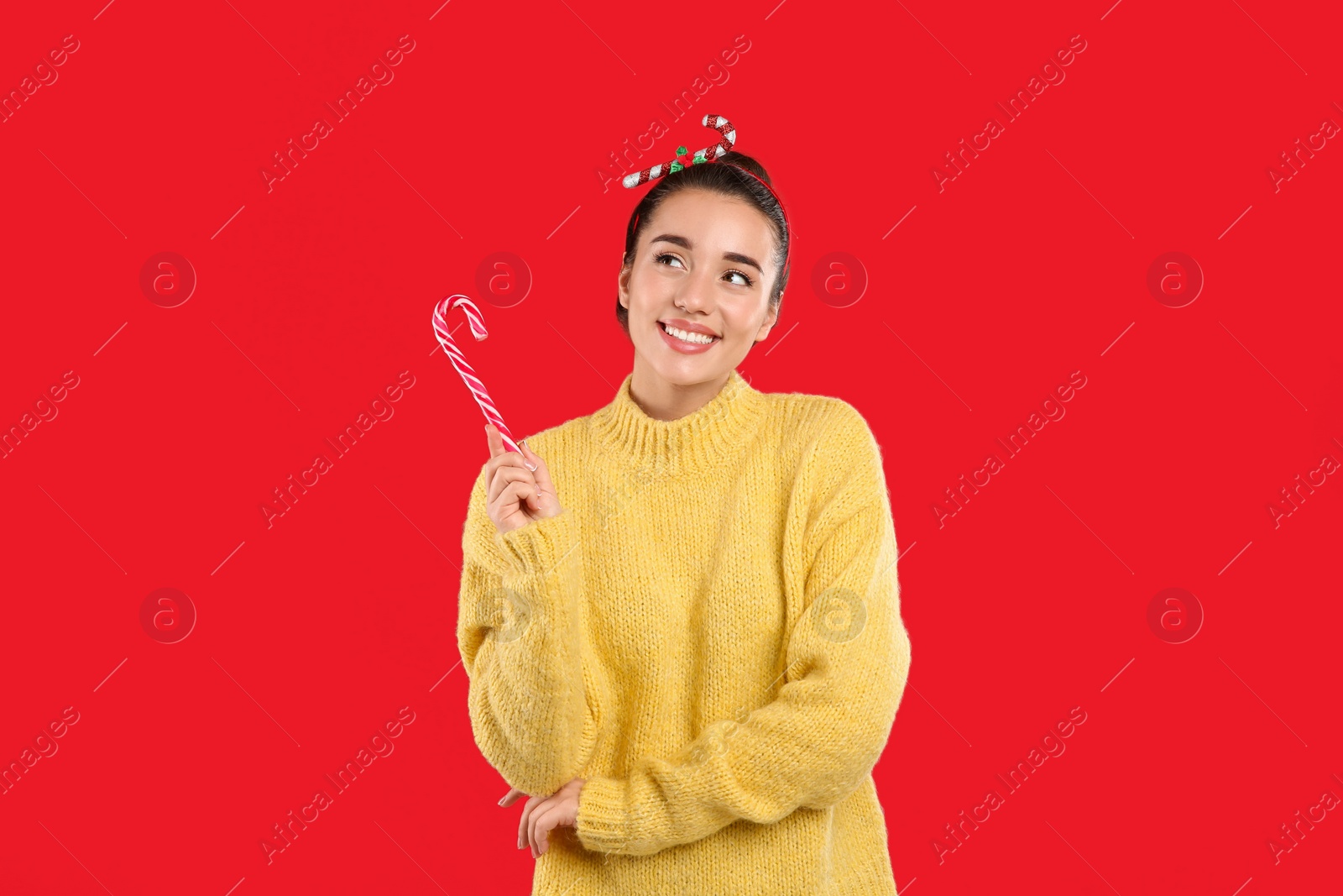 Young woman in yellow sweater and festive headband holding candy cane on red background Photo of Young woman in yellow sweater and festive headband holding candy cane on red background