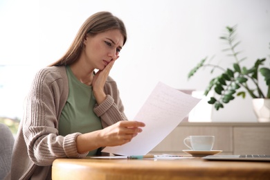 Photo of Worried woman reading letter at wooden table in room