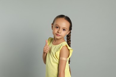 Vaccinated little girl with medical plaster on her arm showing thumb up against light grey background Photo of Vaccinated little girl with medical plaster on her arm showing thumb up against light grey background