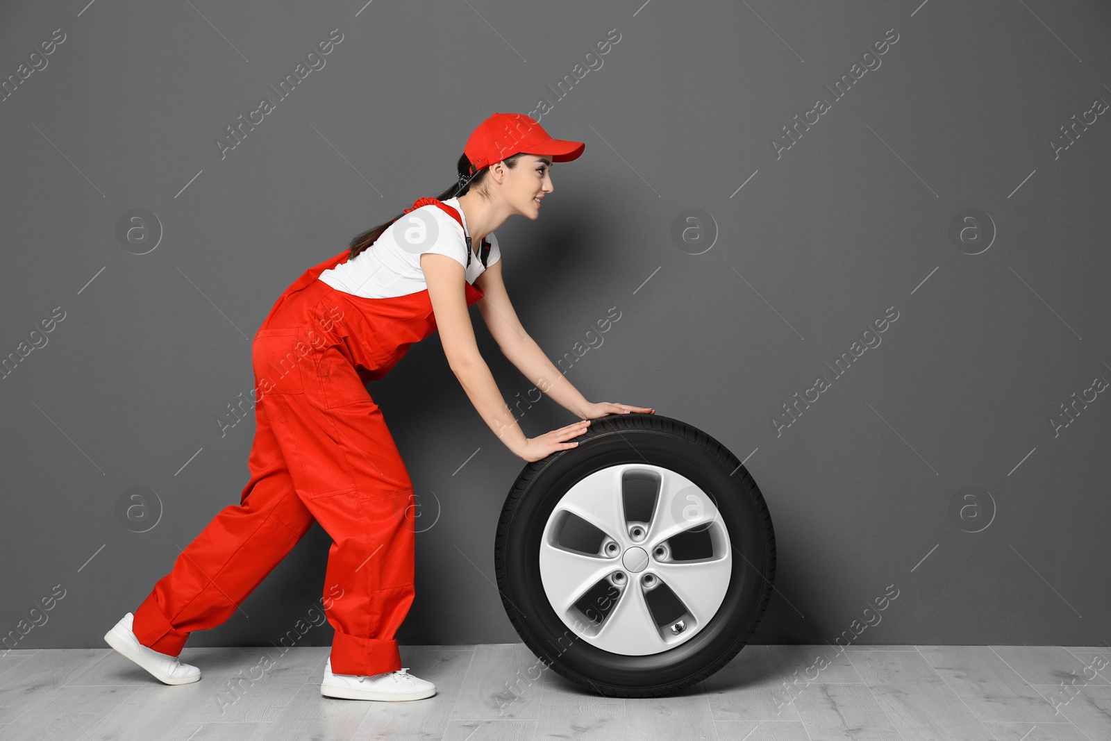 Female mechanic in uniform with car tire on grey wall background Photo of Female mechanic in uniform with car tire on grey wall background