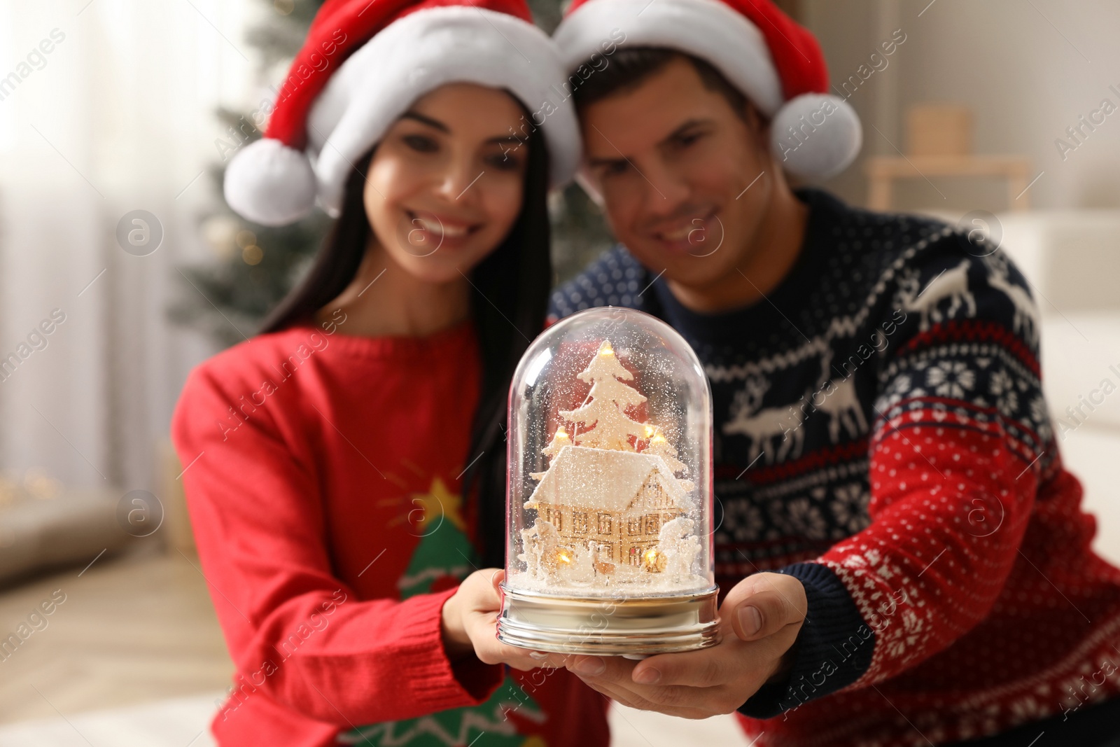Photo of Couple in Santa hats holding snow globe at home
