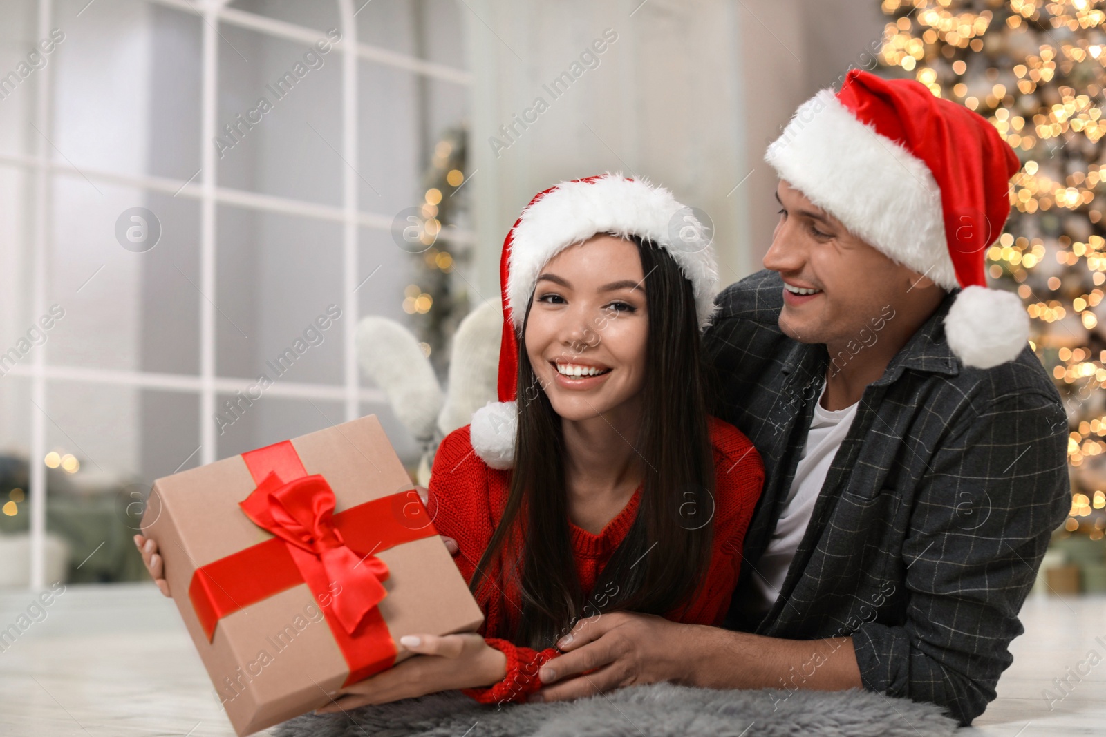 Happy young couple in Santa hats with Christmas gift at home Photo of Happy young couple in Santa hats with Christmas gift at home