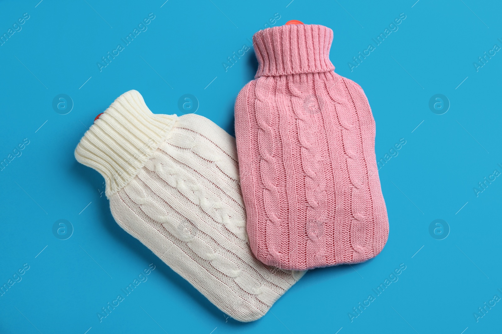 Photo of Hot water bottles with knitted covers on light blue background, flat lay