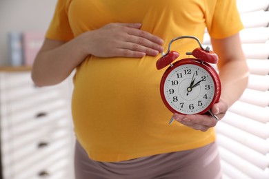 Young pregnant woman holding alarm clock near her belly at home, closeup. Time to give birth Photo of Young pregnant woman holding alarm clock near her belly at home, closeup. Time to give birth