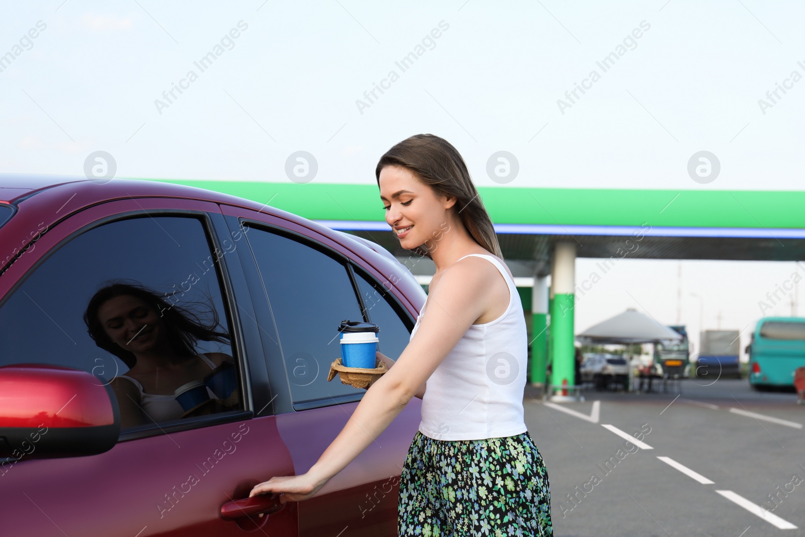Beautiful young woman with coffee opening car door at gas station Photo of Beautiful young woman with coffee opening car door at gas station
