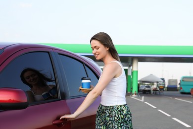 Beautiful young woman with coffee opening car door at gas station Photo of Beautiful young woman with coffee opening car door at gas station