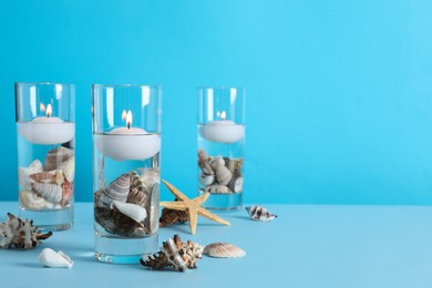 Burning candles in glass holders with seashells on light blue background, space for text Photo of Burning candles in glass holders with seashells on light blue background, space for text