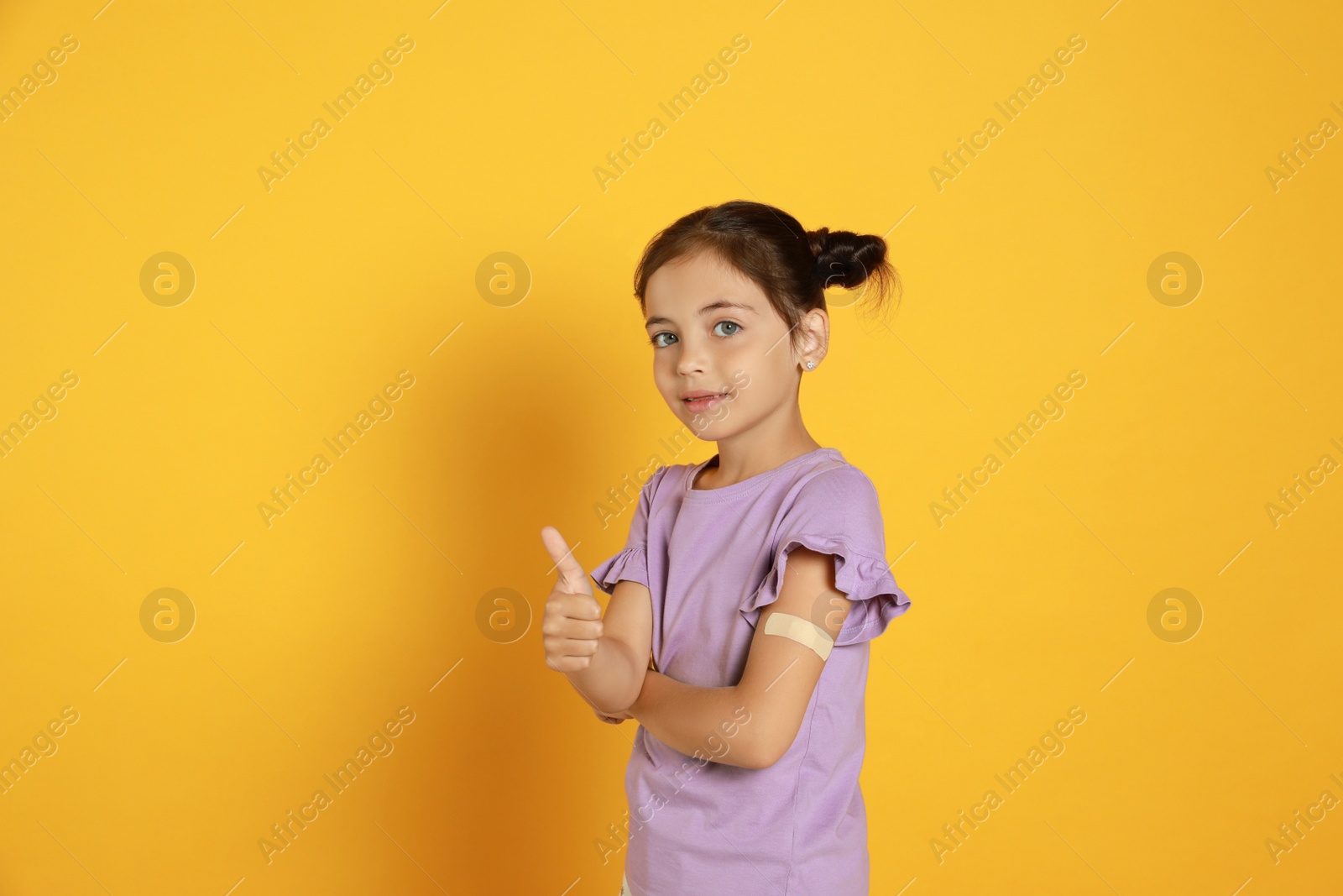 Vaccinated little girl with medical plaster on her arm showing thumb up against yellow background Photo of Vaccinated little girl with medical plaster on her arm showing thumb up against yellow background