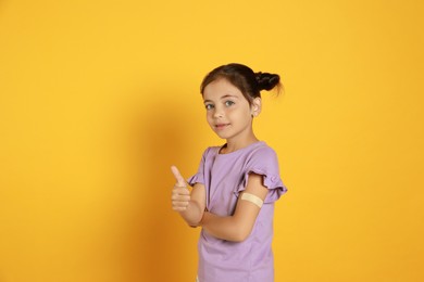 Vaccinated little girl with medical plaster on her arm showing thumb up against yellow background Photo of Vaccinated little girl with medical plaster on her arm showing thumb up against yellow background