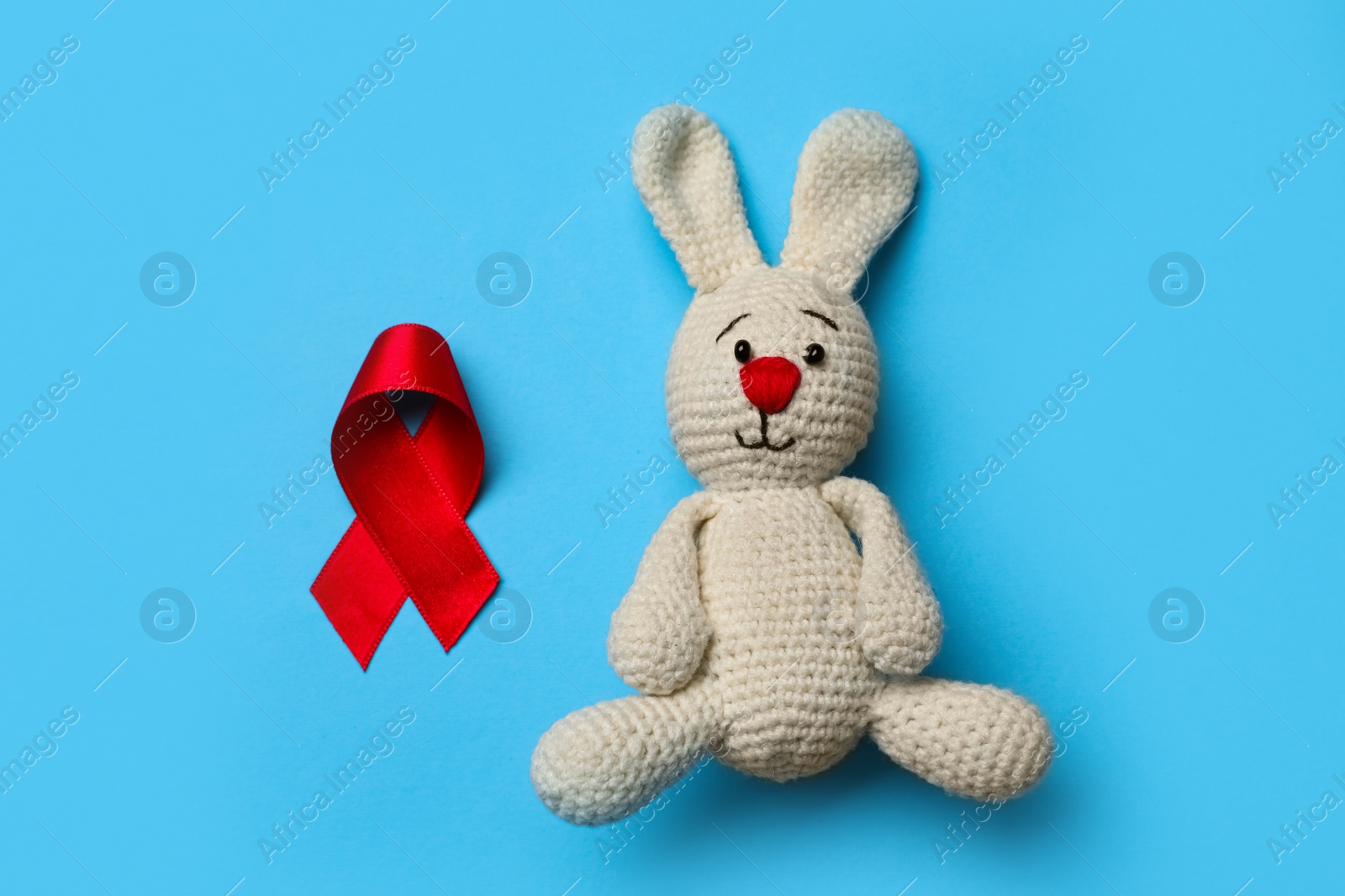 Cute knitted toy bunny and red ribbon on blue background, flat lay. AIDS disease awareness Photo of Cute knitted toy bunny and red ribbon on blue background, flat lay. AIDS disease awareness