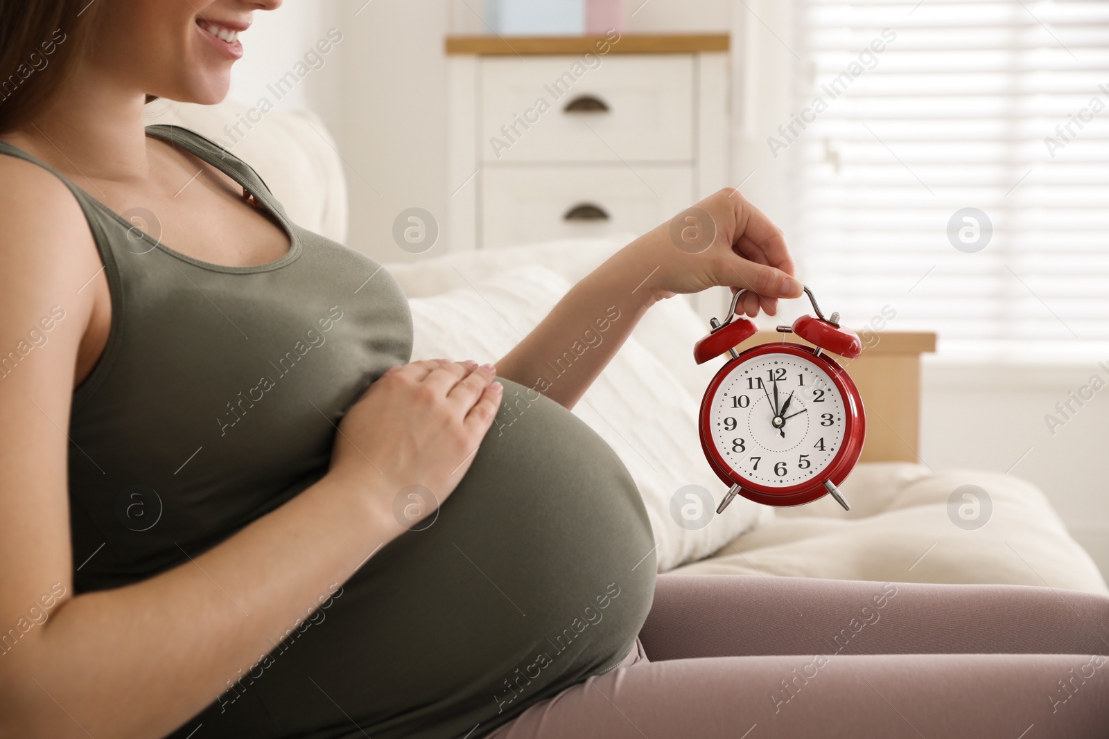 Young pregnant woman holding alarm clock near her belly at home, closeup. Time to give birth Photo of Young pregnant woman holding alarm clock near her belly at home, closeup. Time to give birth