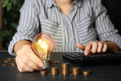 Woman with light bulb, calculator and coins at black wooden table, closeup. Power saving Photo of Woman with light bulb, calculator and coins at black wooden table, closeup. Power saving