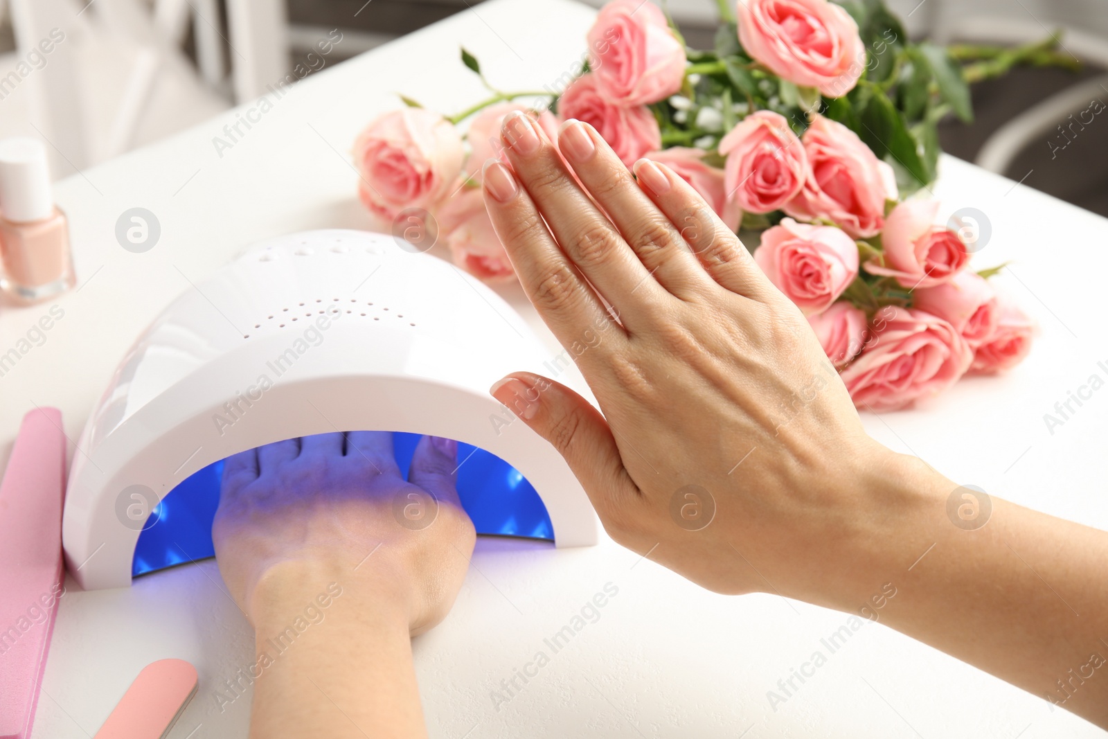 Photo of Woman using ultraviolet lamp to dry gel nail polish at white table, closeup