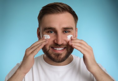 Happy young man applying facial cream on light blue background Photo of Happy young man applying facial cream on light blue background