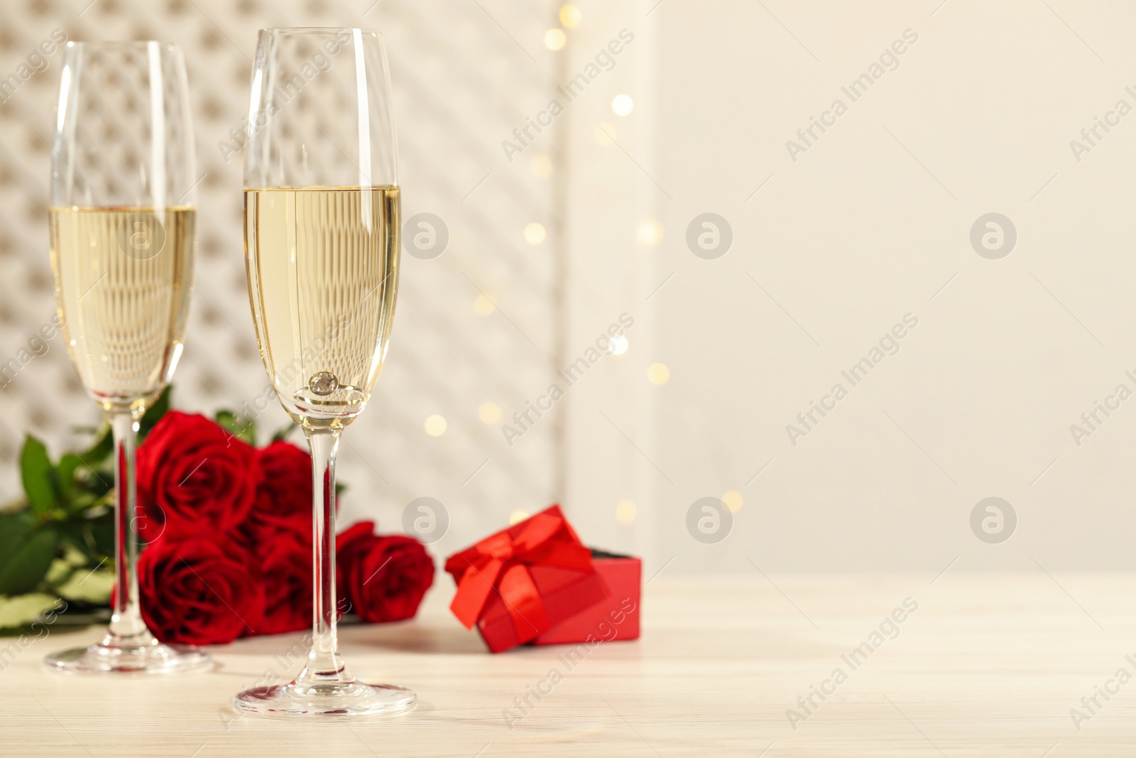 Beautiful engagement ring with gemstone in glass of sparkling wine and roses on white wooden table. Space for text Photo of Beautiful engagement ring with gemstone in glass of sparkling wine and roses on white wooden table. Space for text