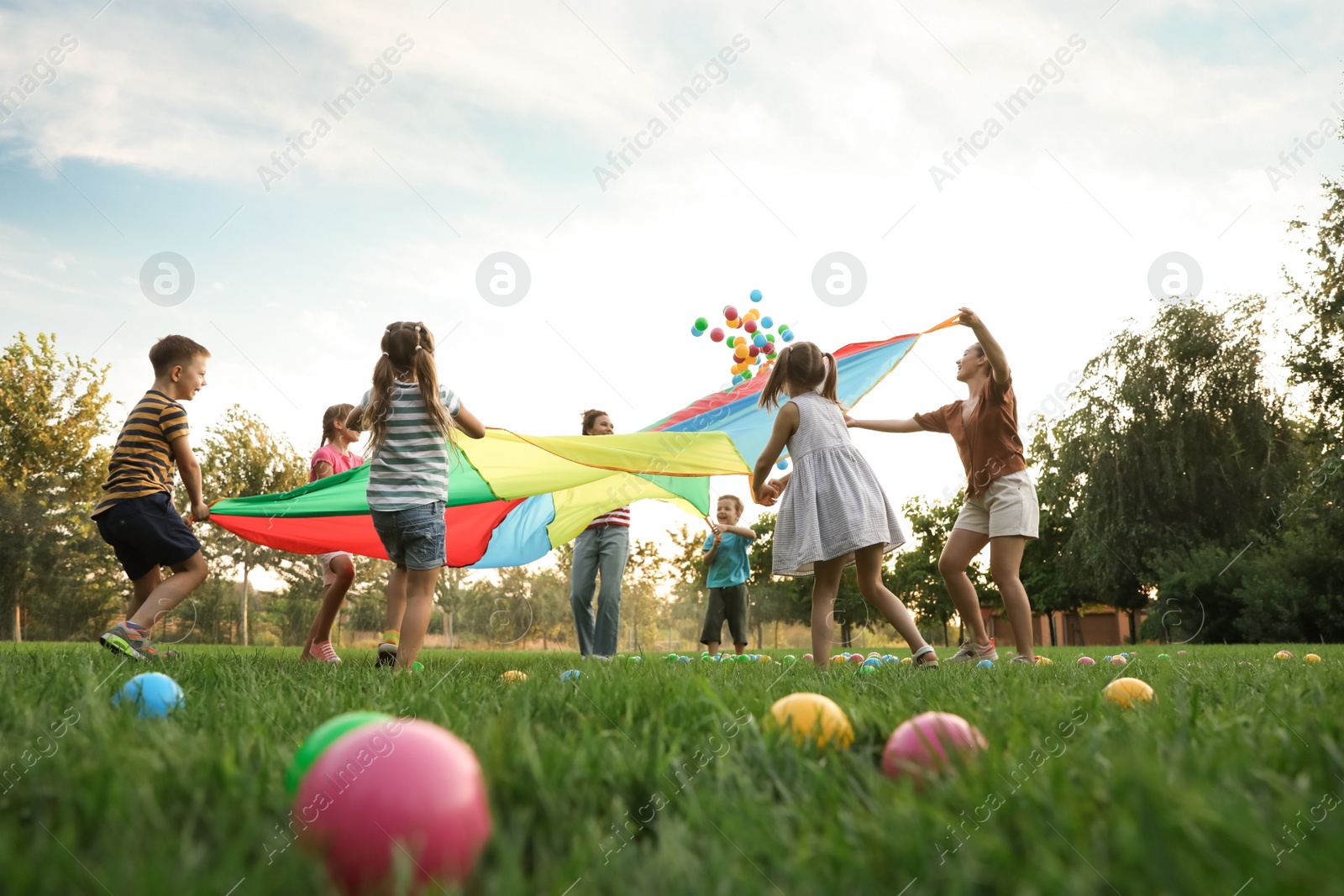 Photo of Group of children and teachers playing with rainbow playground parachute on green grass, low angle view. Summer camp activity