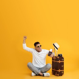 Emotional male tourist holding passport with ticket near suitcase on yellow background Photo of Emotional male tourist holding passport with ticket near suitcase on yellow background