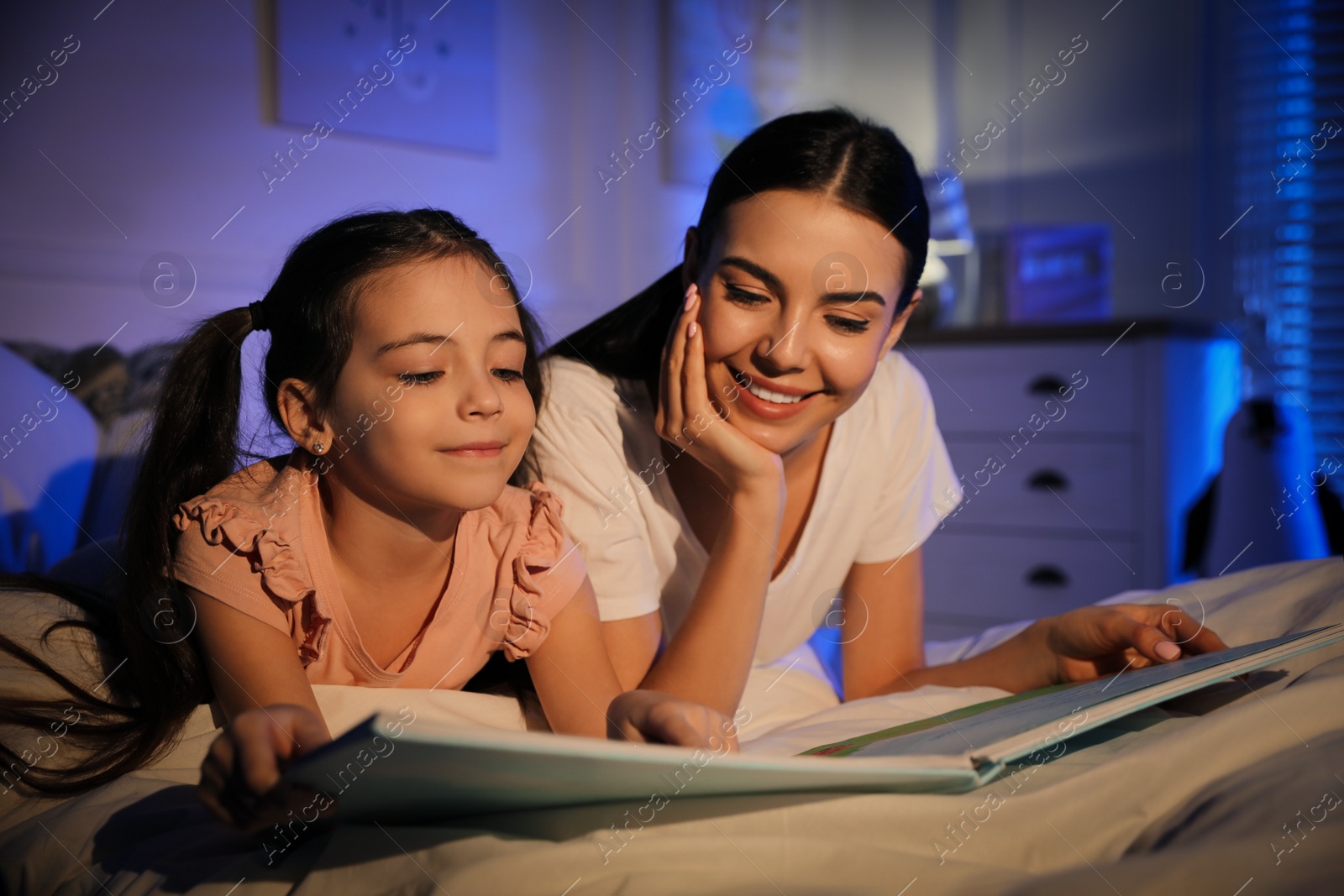 Photo of Mother with little daughter reading fairy tale in dark bedroom
