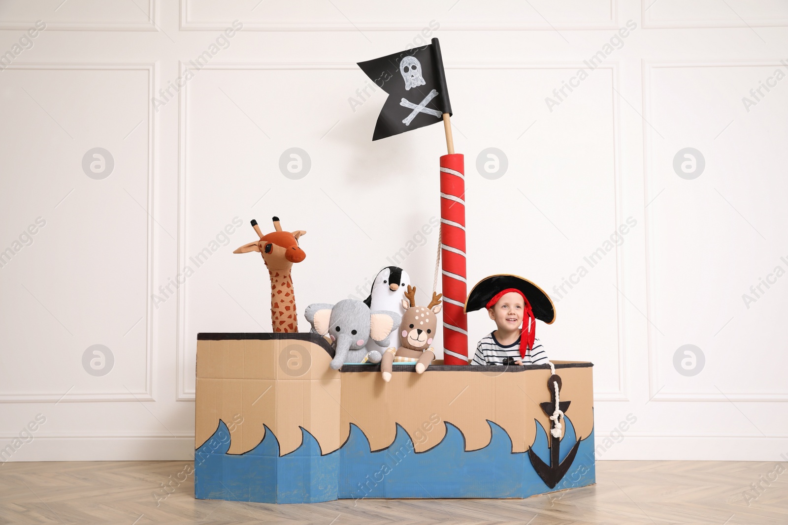 Cute little boy playing with toys in pirate cardboard ship near white wall indoors Photo of Cute little boy playing with toys in pirate cardboard ship near white wall indoors