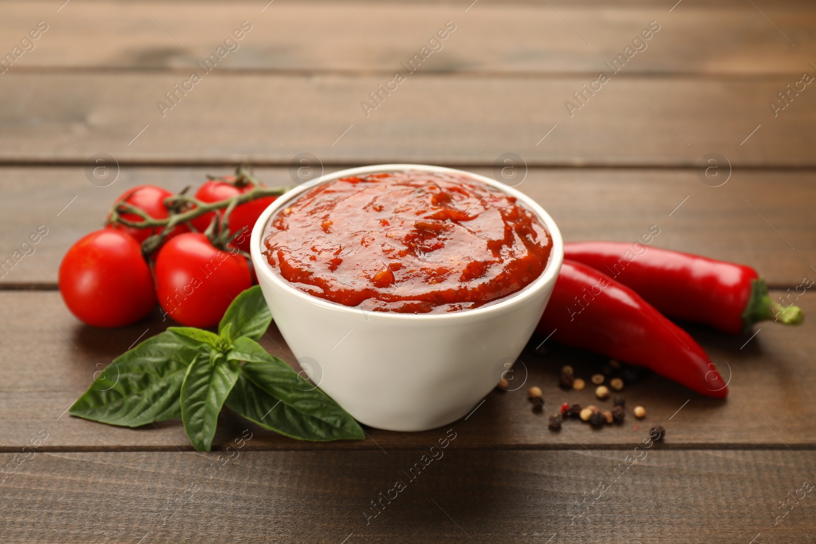 Delicious adjika sauce in bowl and ingredients on wooden table Photo of Delicious adjika sauce in bowl and ingredients on wooden table