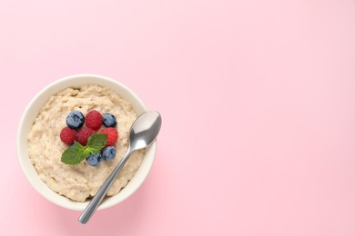 Photo of Tasty oatmeal porridge with raspberries, blueberries and spoon in bowl on pink background, top view