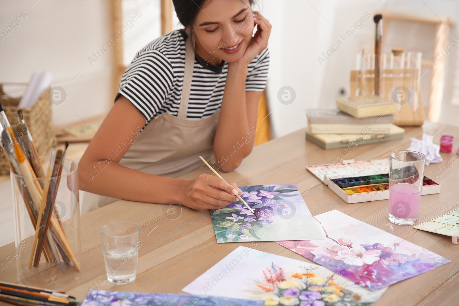 Young woman drawing flowers with watercolors at table indoors, focus on painting Photo of Young woman drawing flowers with watercolors at table indoors, focus on painting