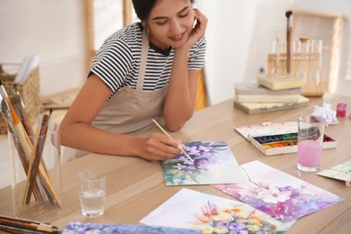 Young woman drawing flowers with watercolors at table indoors, focus on painting Photo of Young woman drawing flowers with watercolors at table indoors, focus on painting