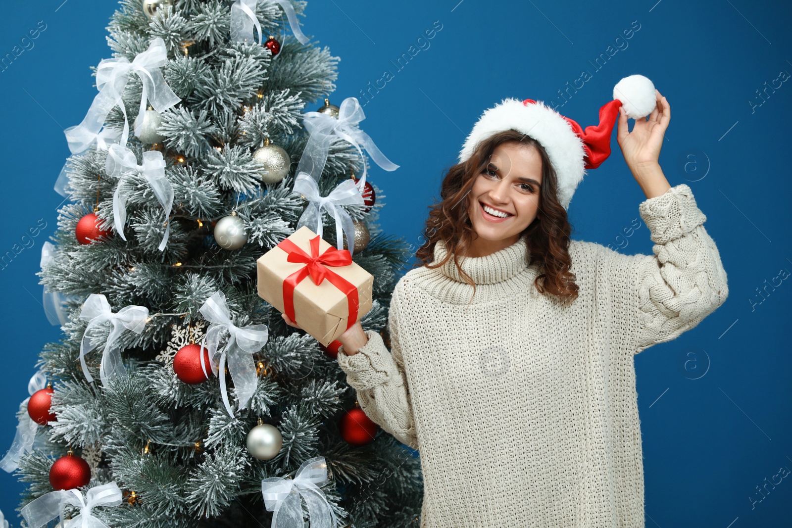 Happy young woman in Santa hat with gift near Christmas tree on blue background Image of Happy young woman in Santa hat with gift near Christmas tree on blue background