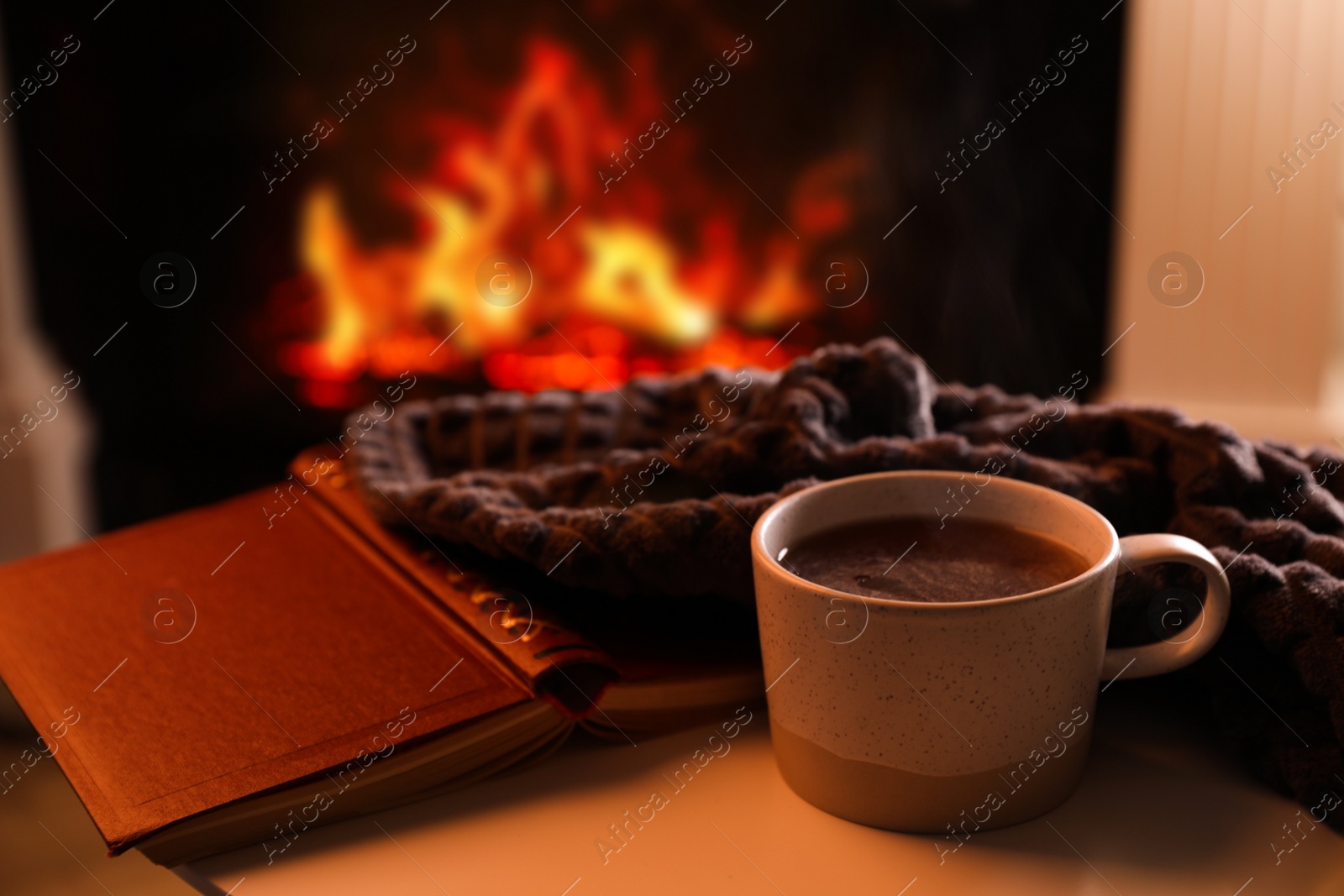 Cup of hot drink and book on table near fireplace indoors Photo of Cup of hot drink and book on table near fireplace indoors