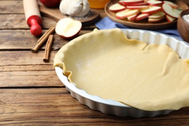 Baking dish with raw dough for apple pie and ingredients on wooden table Photo of Baking dish with raw dough for apple pie and ingredients on wooden table