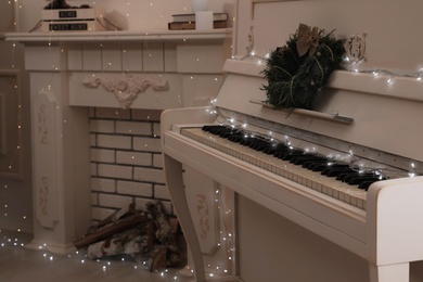 White piano with fairy lights and wreath near decorative fireplace indoors. Christmas music Photo of White piano with fairy lights and wreath near decorative fireplace indoors. Christmas music