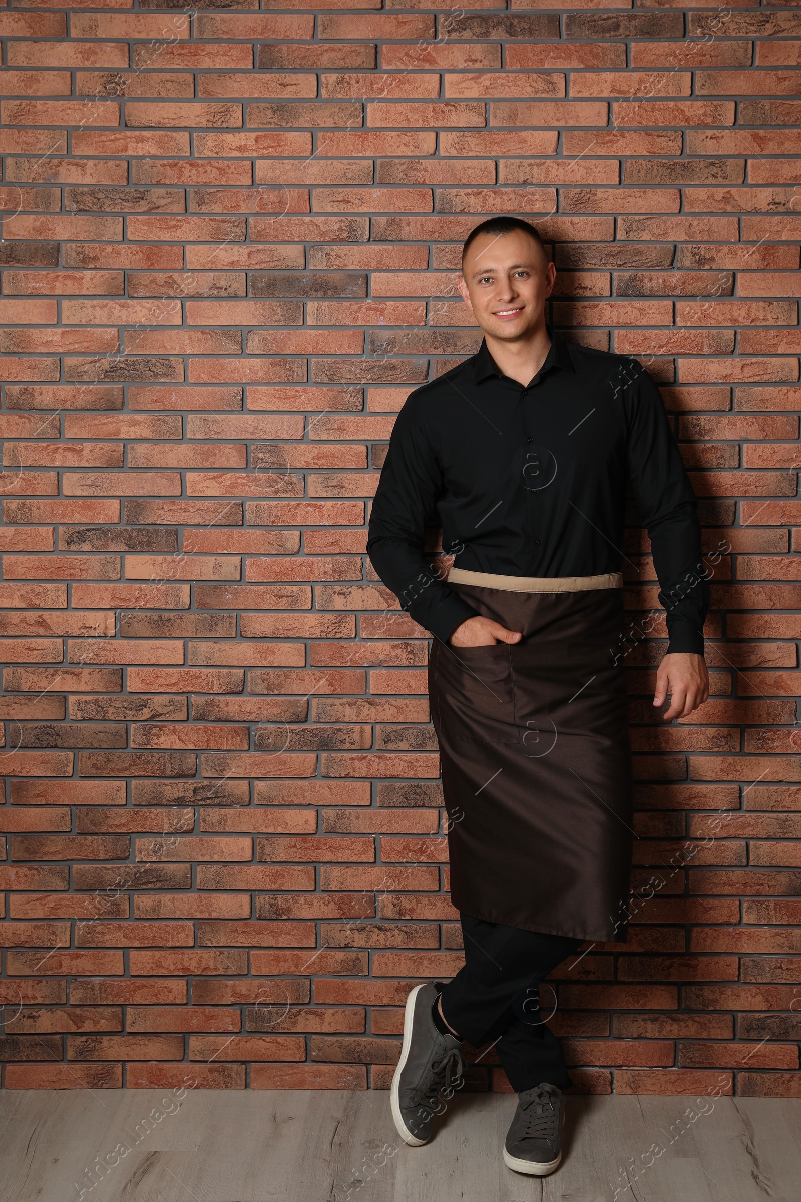 Full length portrait of happy young waiter in uniform near brick wall indoors Photo of Full length portrait of happy young waiter in uniform near brick wall indoors