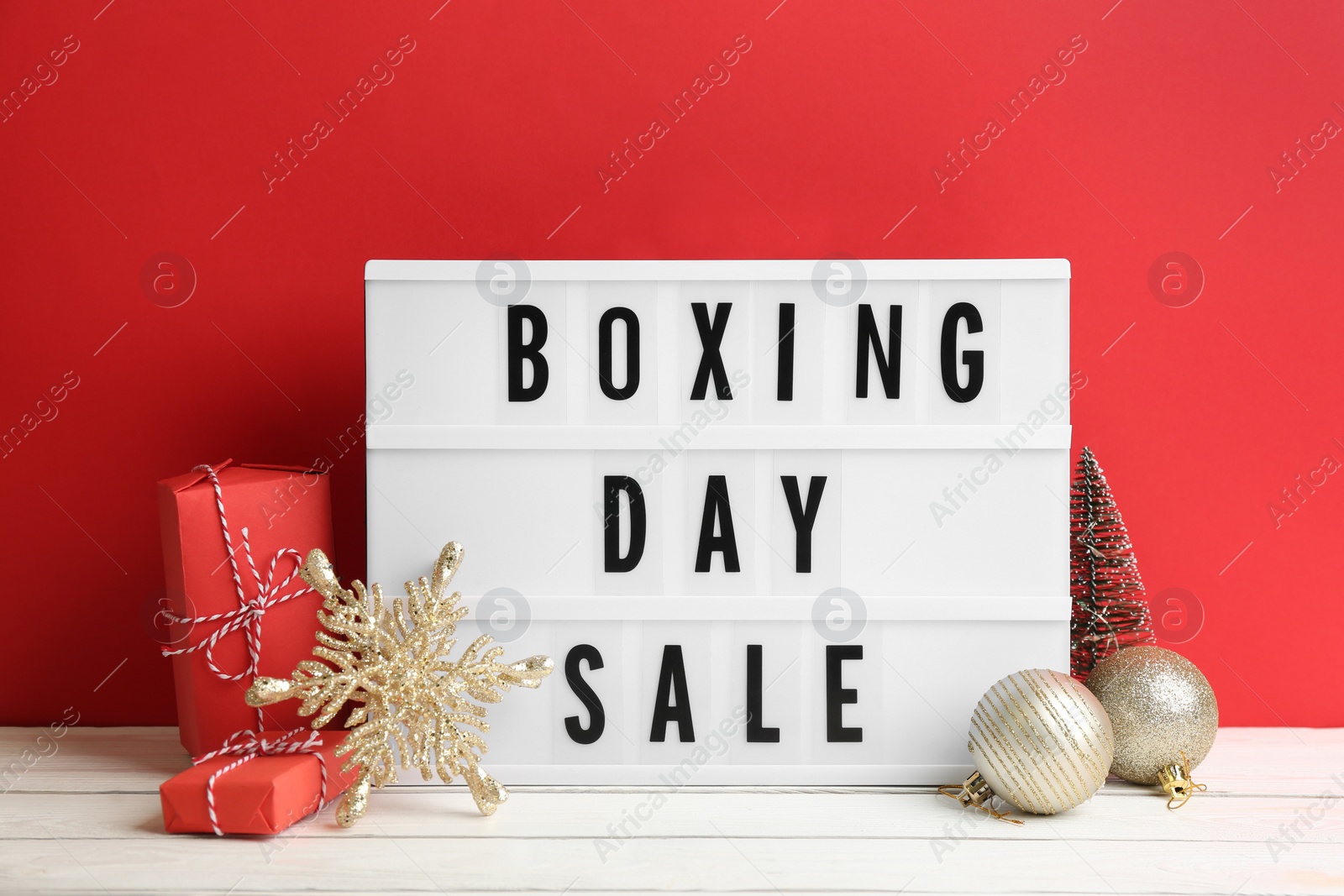 Composition with Boxing Day Sale sign and Christmas gifts on white table against red background Photo of Composition with Boxing Day Sale sign and Christmas gifts on white table against red background