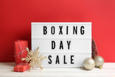 Composition with Boxing Day Sale sign and Christmas gifts on white table against red background Photo of Composition with Boxing Day Sale sign and Christmas gifts on white table against red background