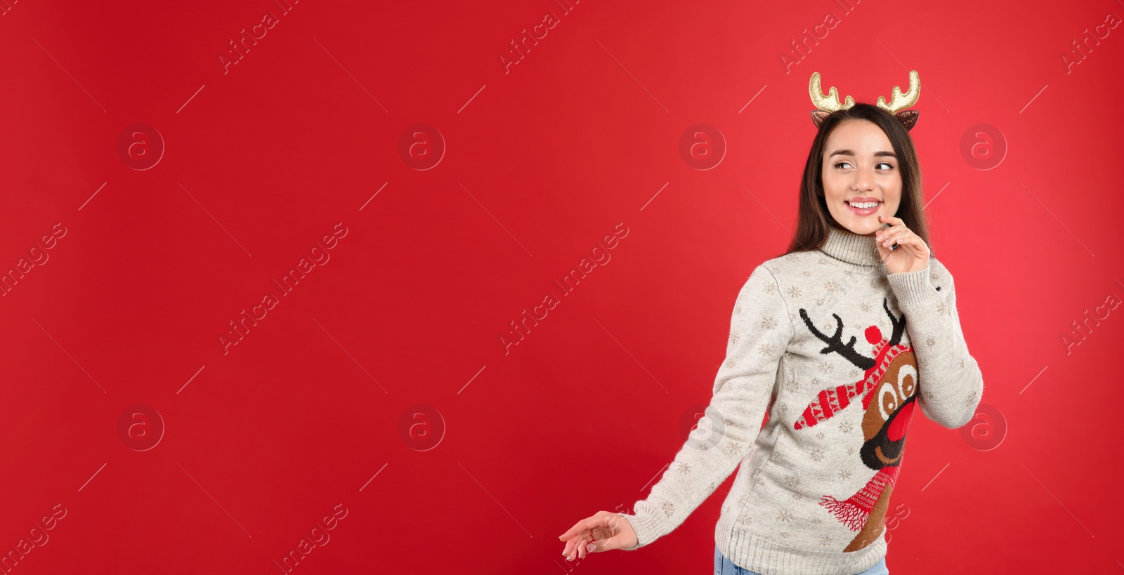 Young woman in Christmas sweater and deer headband on red background, space for text Photo of Young woman in Christmas sweater and deer headband on red background, space for text