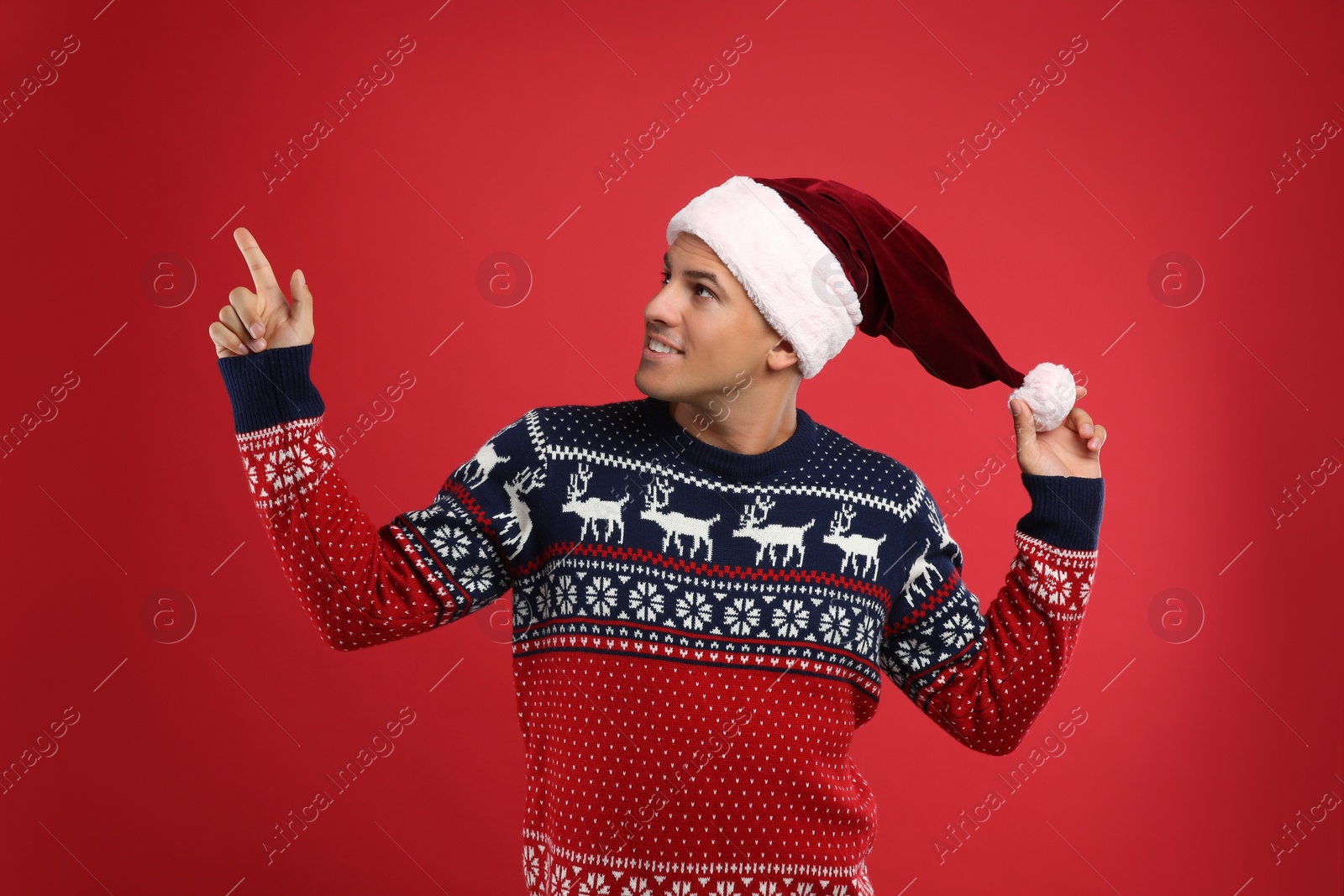 Handsome man in Christmas sweater and Santa hat pointing on red background Photo of Handsome man in Christmas sweater and Santa hat pointing on red background