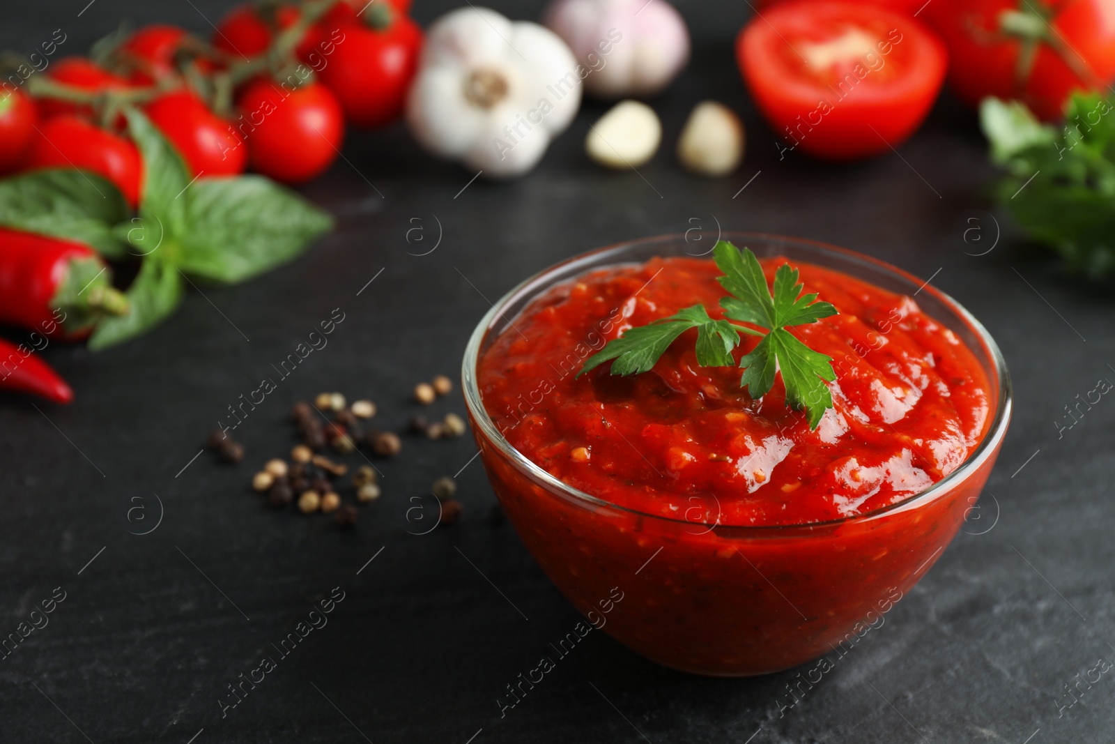 Delicious adjika sauce with parsley in glass bowl and ingredients on black table, closeup Photo of Delicious adjika sauce with parsley in glass bowl and ingredients on black table, closeup